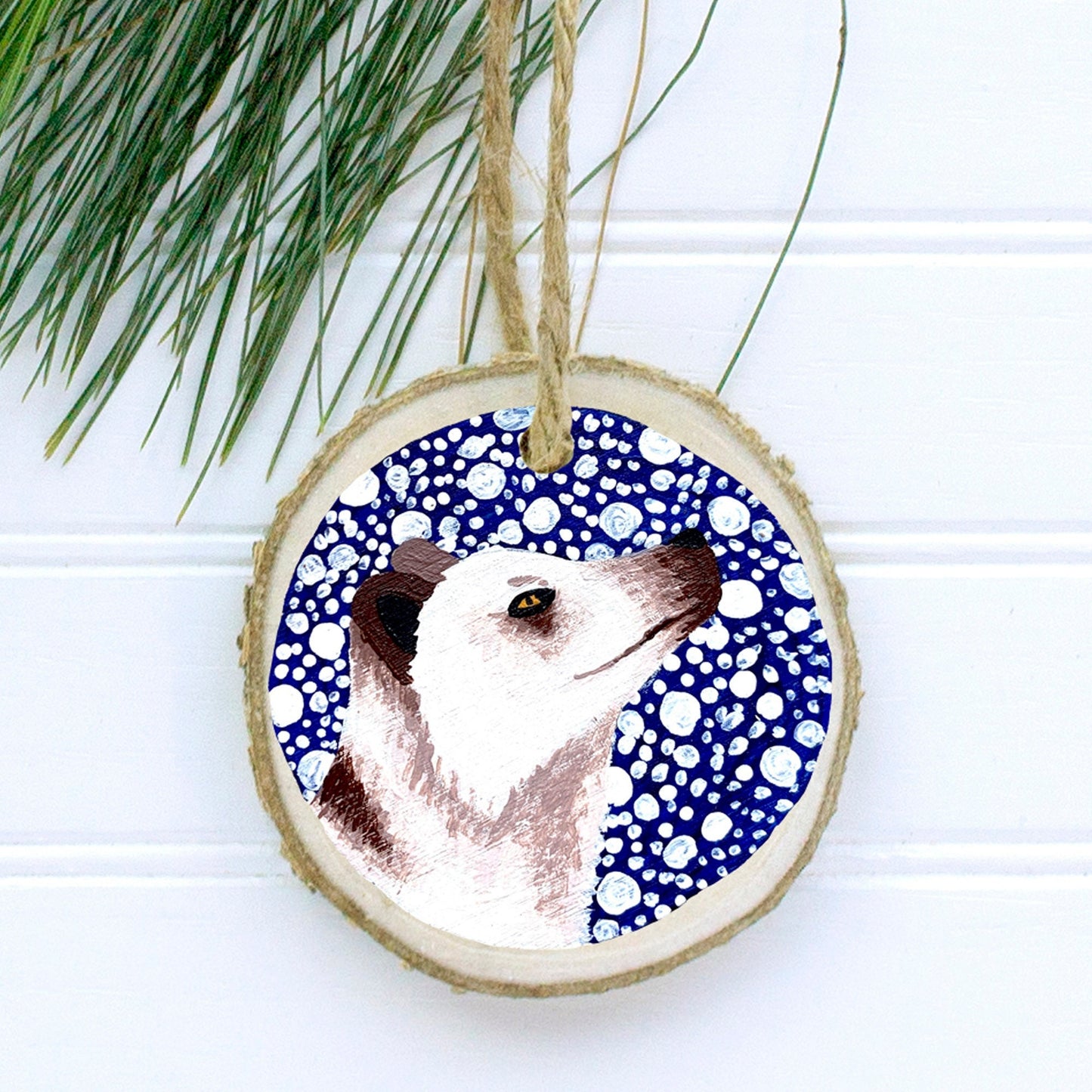 A hand-painted wooden Christmas ornament featuring a white Arctic fox on a dark blue snowy background, with natural pine wood and a jute twine string for hanging.