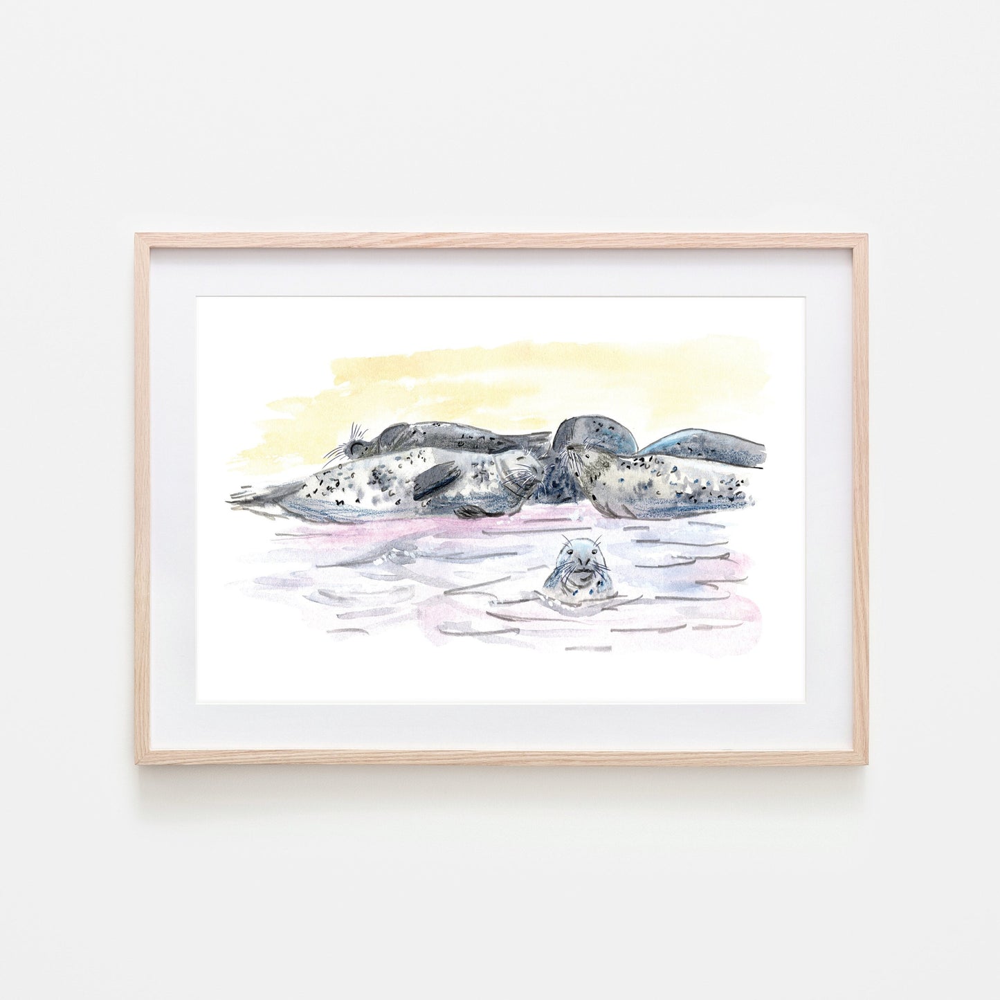 A framed art print depicting a group of grey seals resting on a beach, with watercolour and wax pastel techniques.