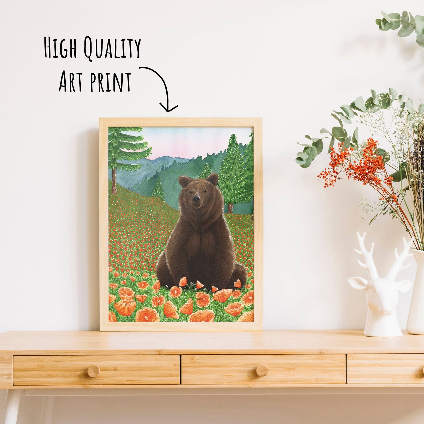 Showing the grizzly bear art print in a light wooden frame sat on a wooden table top with a vase of dried foliage.