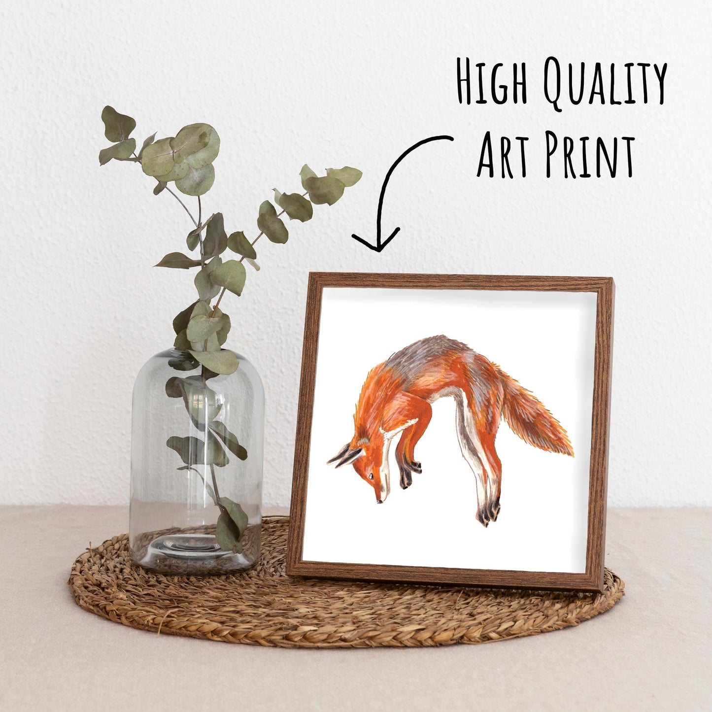 Jumping Red Fox Art Print framed in a dark wood frame on a straw placemat with a vase with a eucalyptus branch.