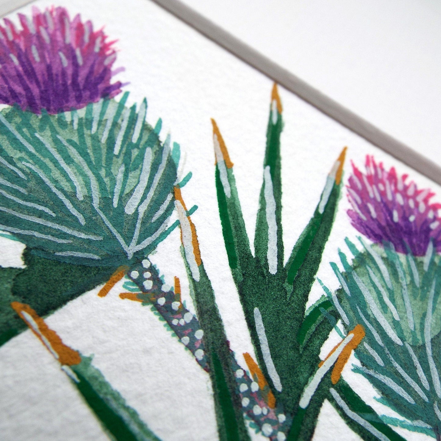 Close-up of a watercolour and gouache thistle painting of green leaves with purple flowers on a white paper background.