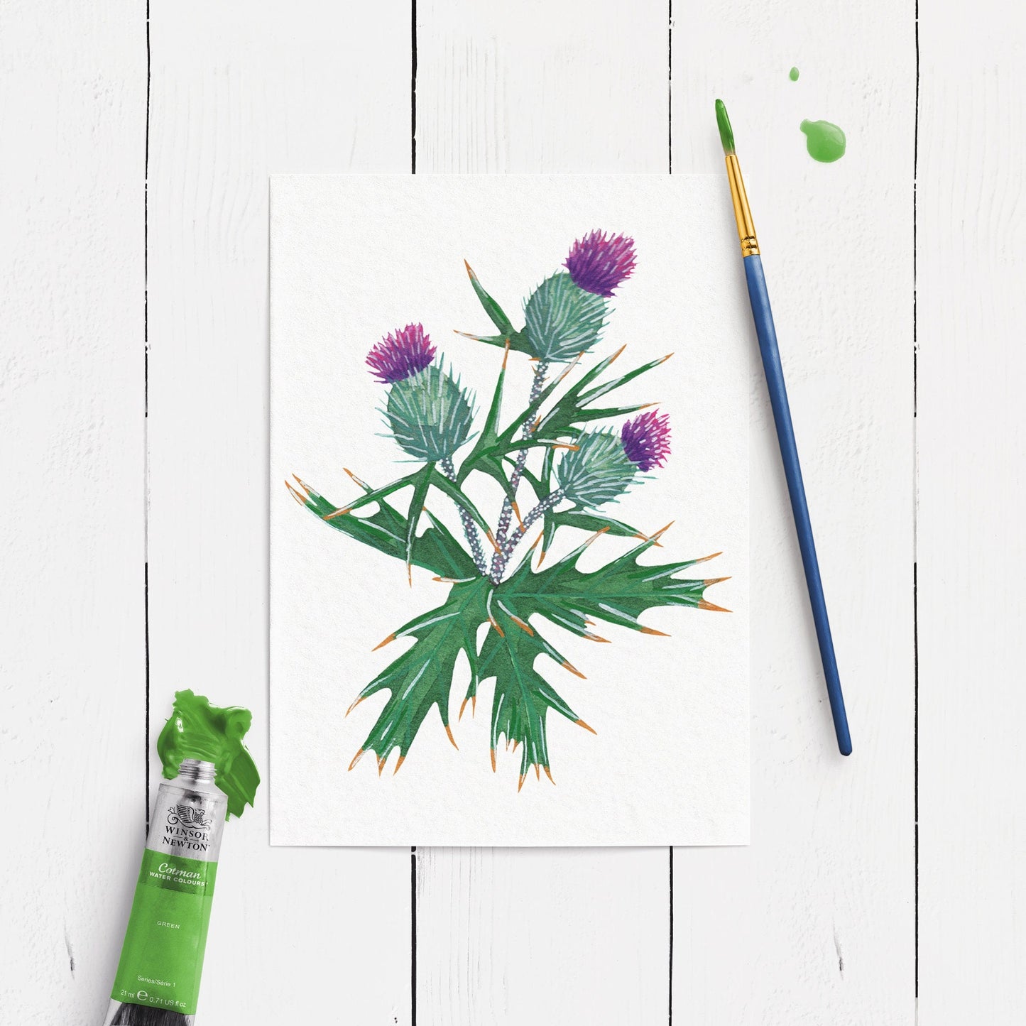 Painting of a thistle plant with a paintbrush and tube of green paint on a white wooden surface.