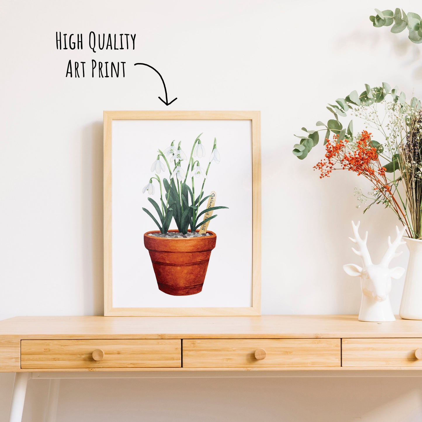 Watercolour snowdrops in a Terracota pot art print in a light wooden framed on a table top with a vase of dried foliage