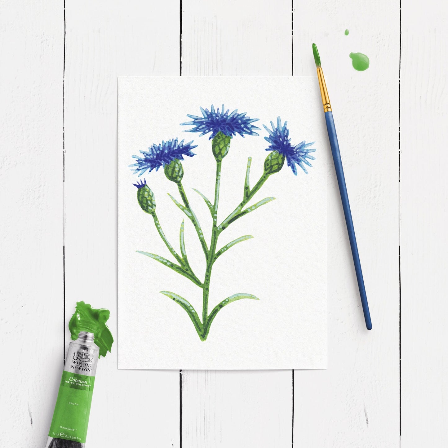 Painting of blue flowers with a paintbrush and paint tube on a white wooden surface