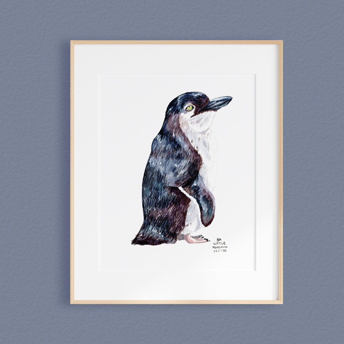 A framed mixed media original painting of a Little Penguin, featuring a light wood frame and white matting, displayed against a textured blue grey wall.