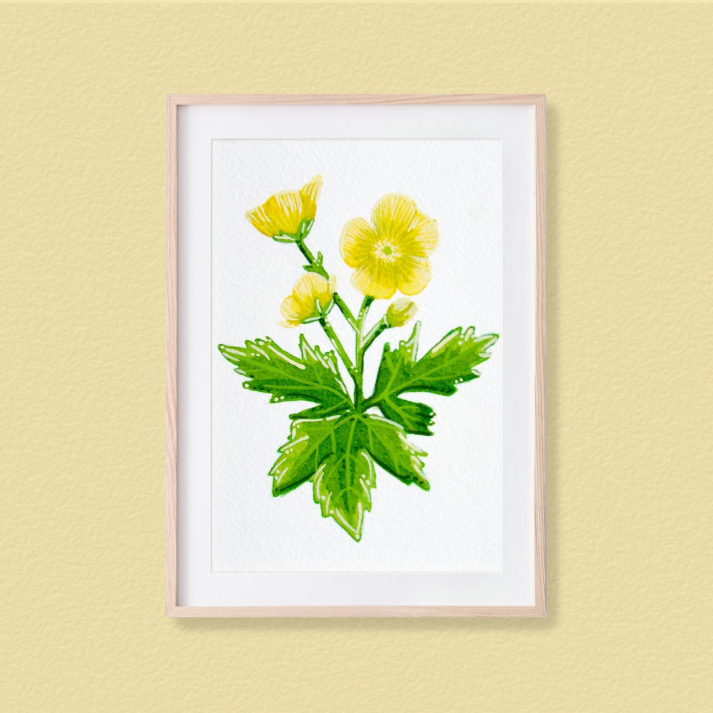 A framed mixed media original painting of a yellow buttercup flower featuring a light wood frame on white background, displayed against a textured pale yellow wall.