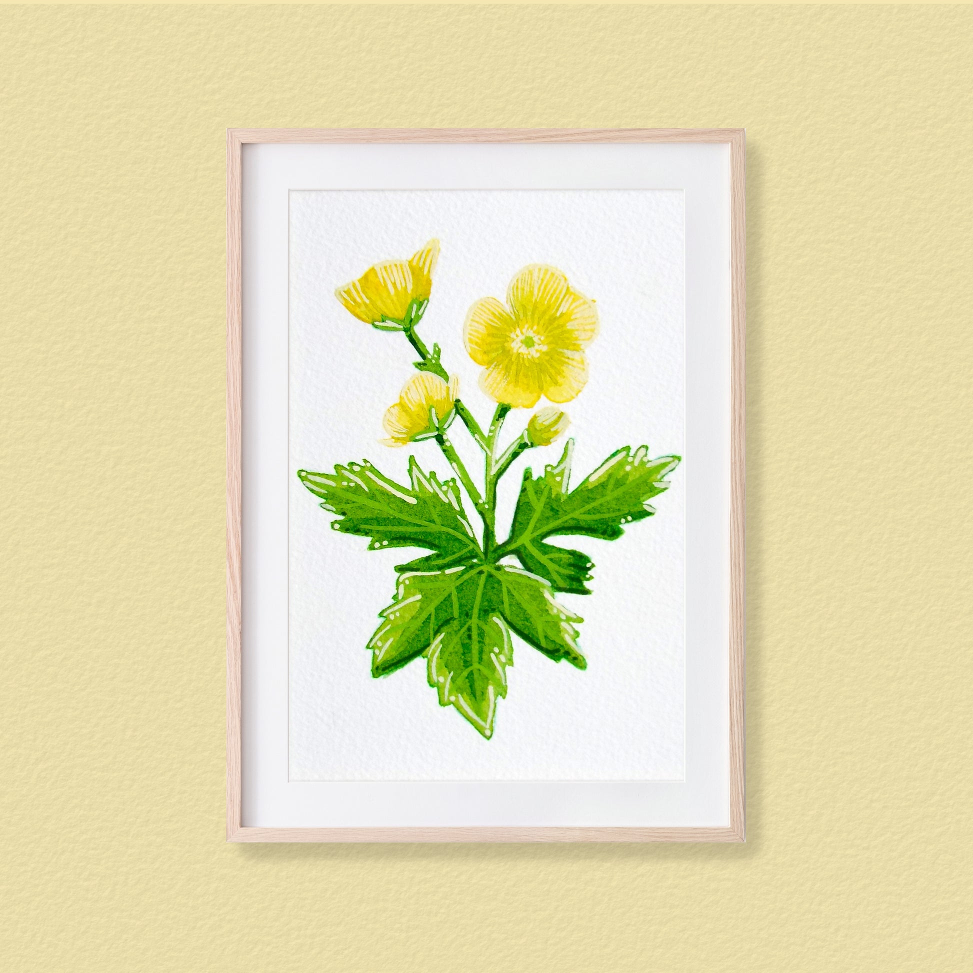 A framed mixed media original painting of a yellow buttercup flower featuring a light wood frame on white background, displayed against a textured pale yellow wall.