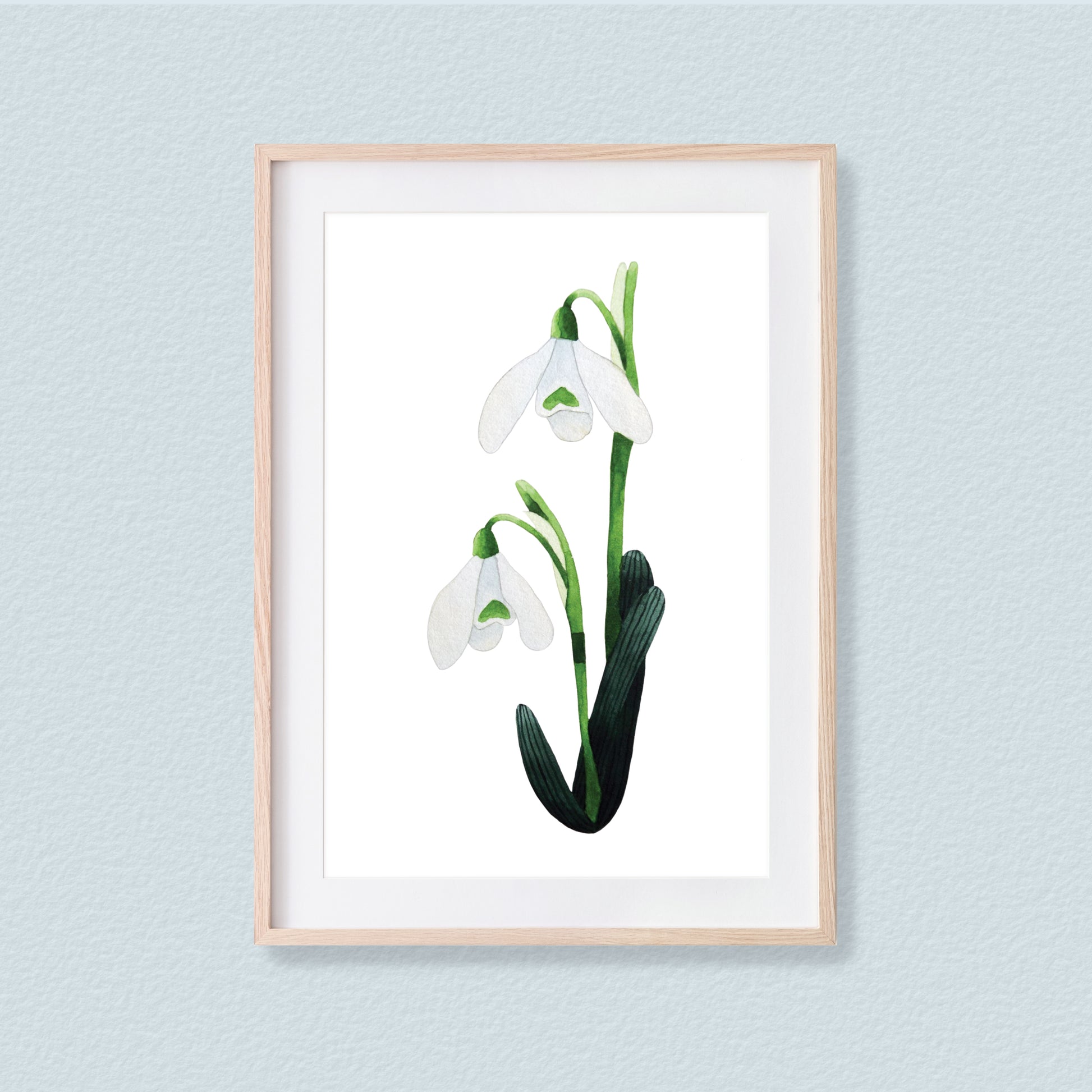 A framed mixed media original painting of a snowdrop flower featuring a light wood frame on white background, displayed against a textured light pale blue wall.