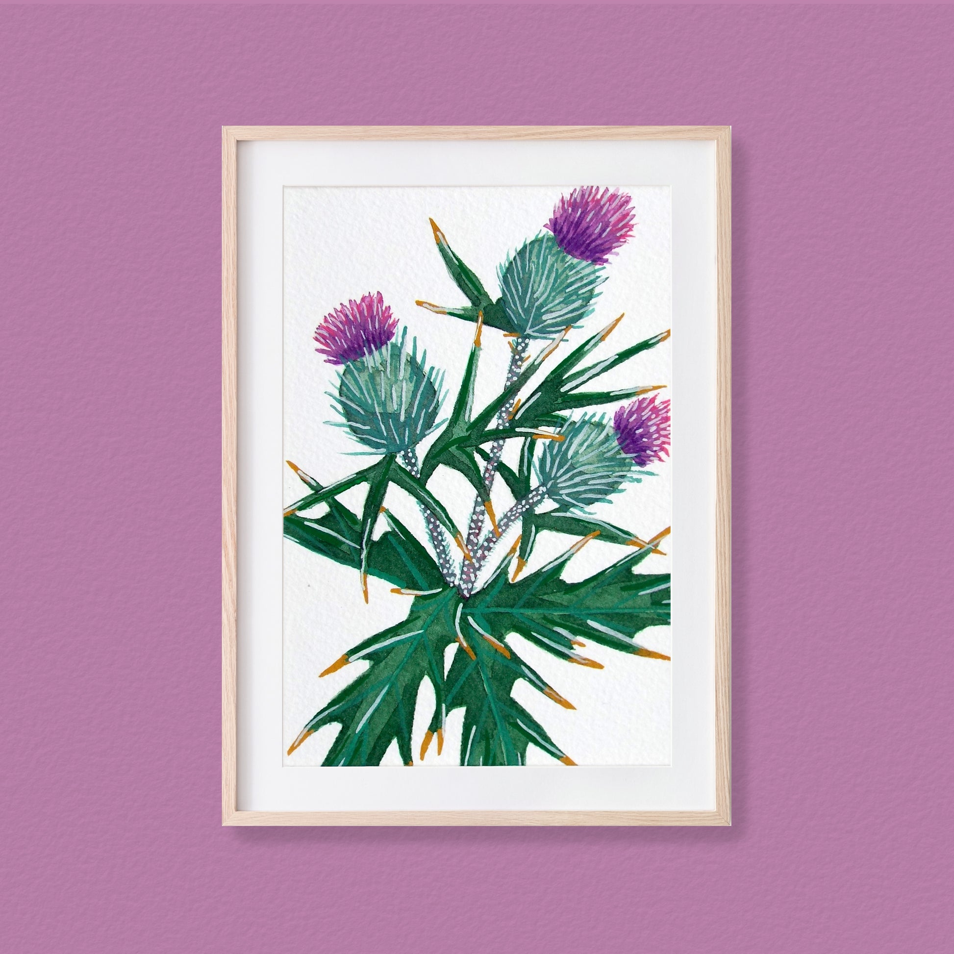 A framed mixed media original painting of a purple thistle flower featuring a light wood frame on white, displayed against a textured purple-pink wall.