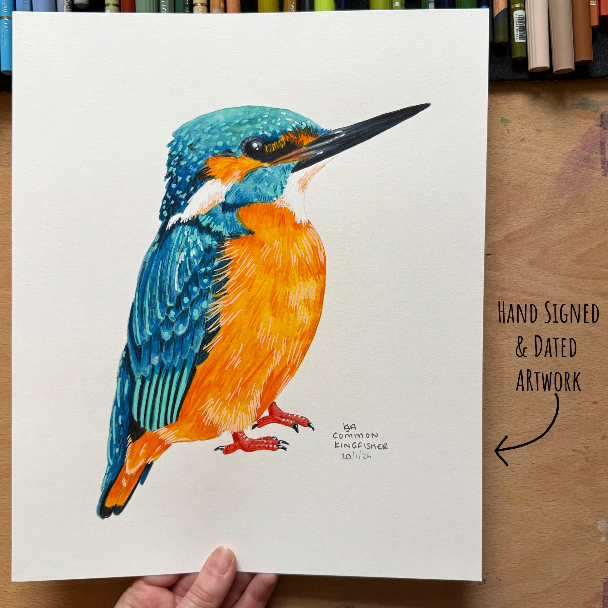 Colourful bird illustration on a white sheet with 'Hand Signed & Dated Artwork' text. being held over a wooden surface featuring art supplies.