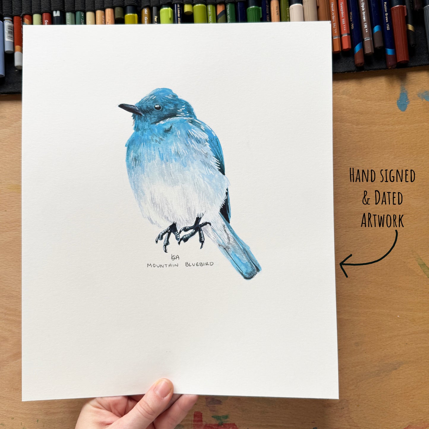Hand-signed and dated artwork of a blue bird on paper, held by a hand on a wooden surface with art supplies.