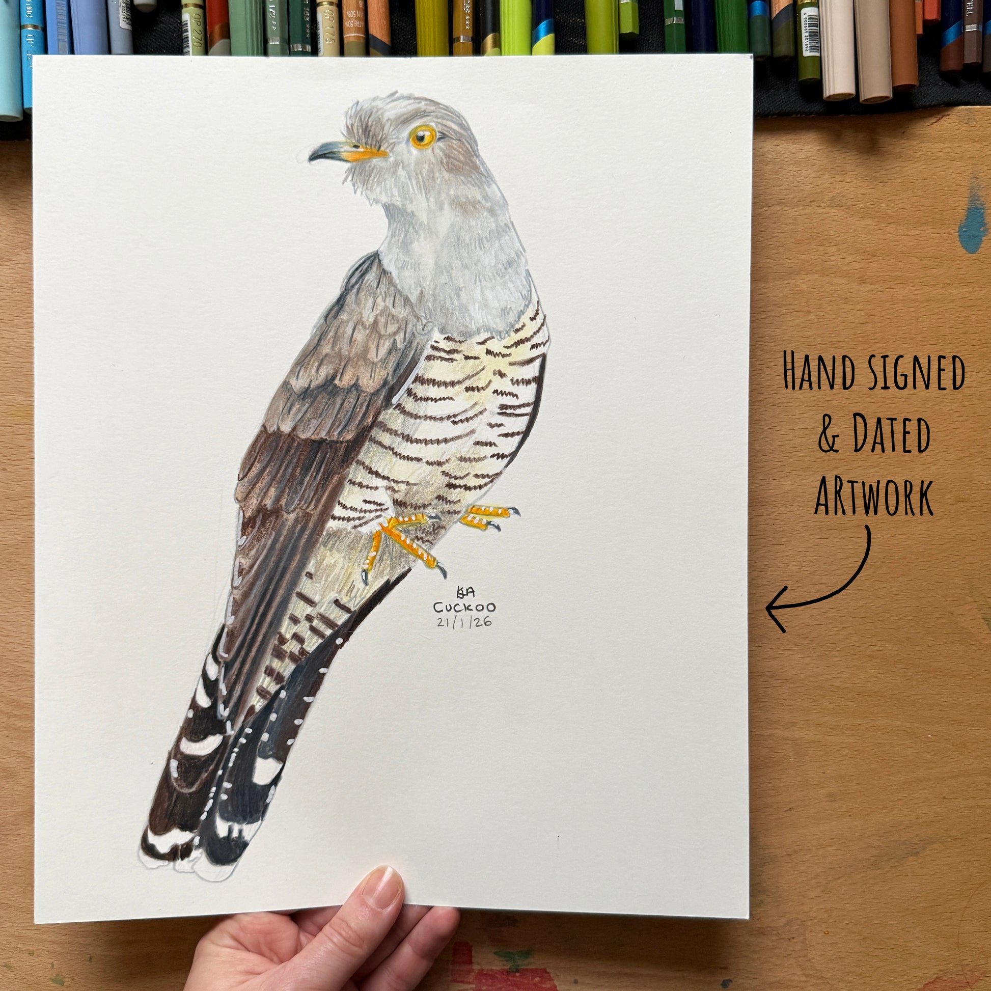 Hand-signed and dated artwork of a bird on a white sheet held by a hand, with a wooden surface and art supplies in the background.