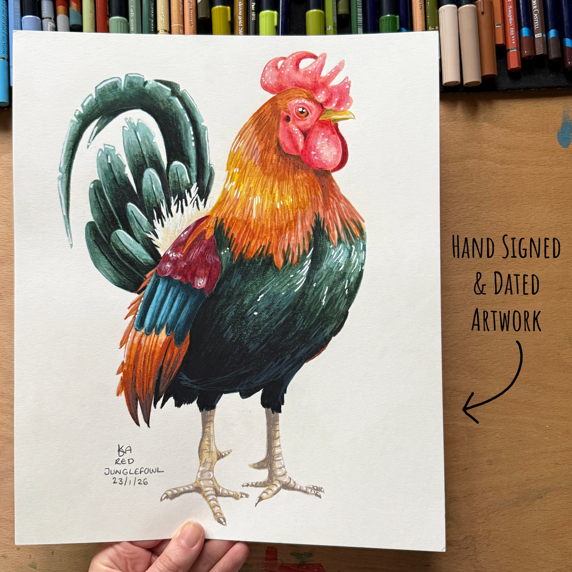 Colourful rooster illustration on a white sheet with a hand holding it, set against a wooden surface with art supplies in the background.