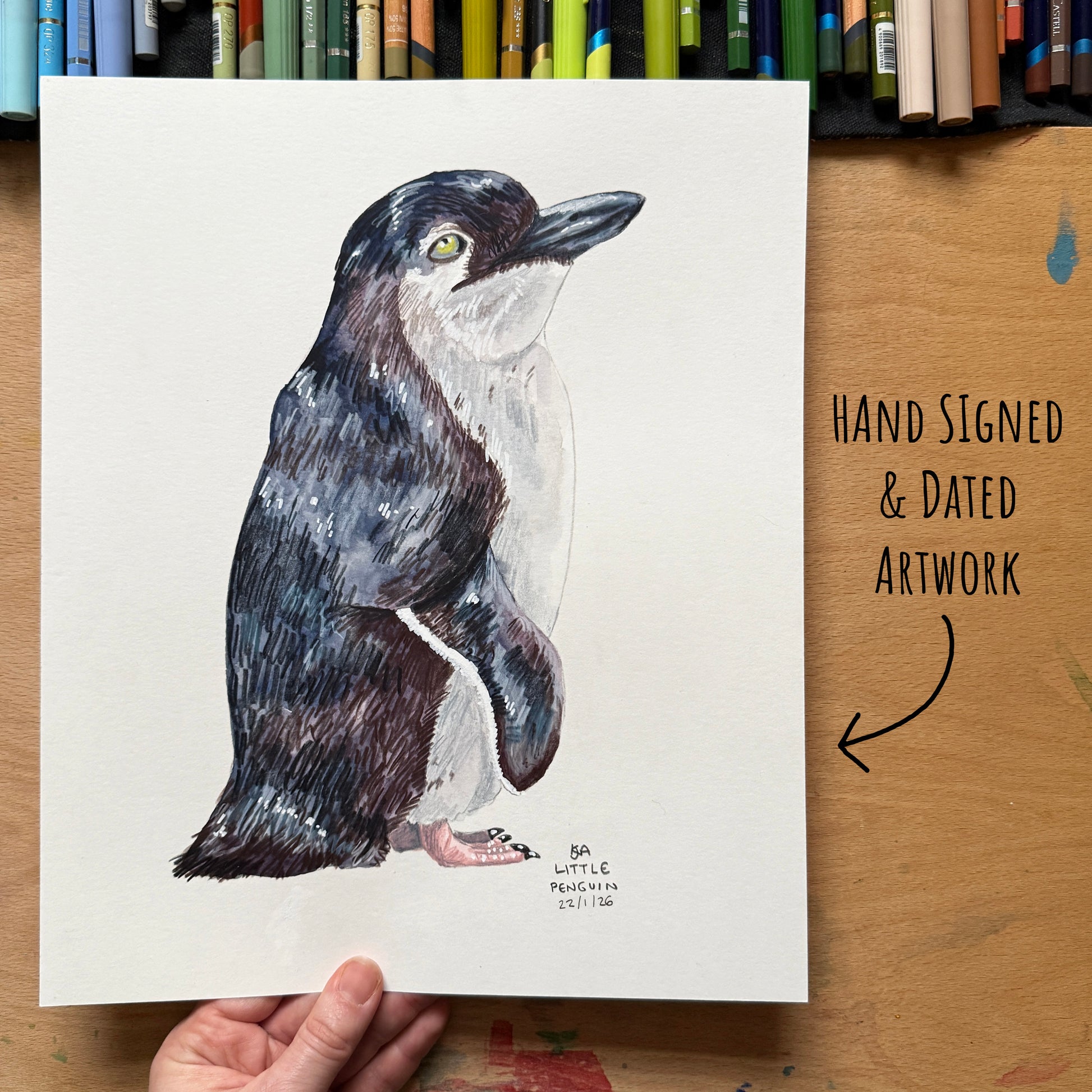 Hand-signed and dated artwork of a penguin on a wooden surface with art supplies in the background.