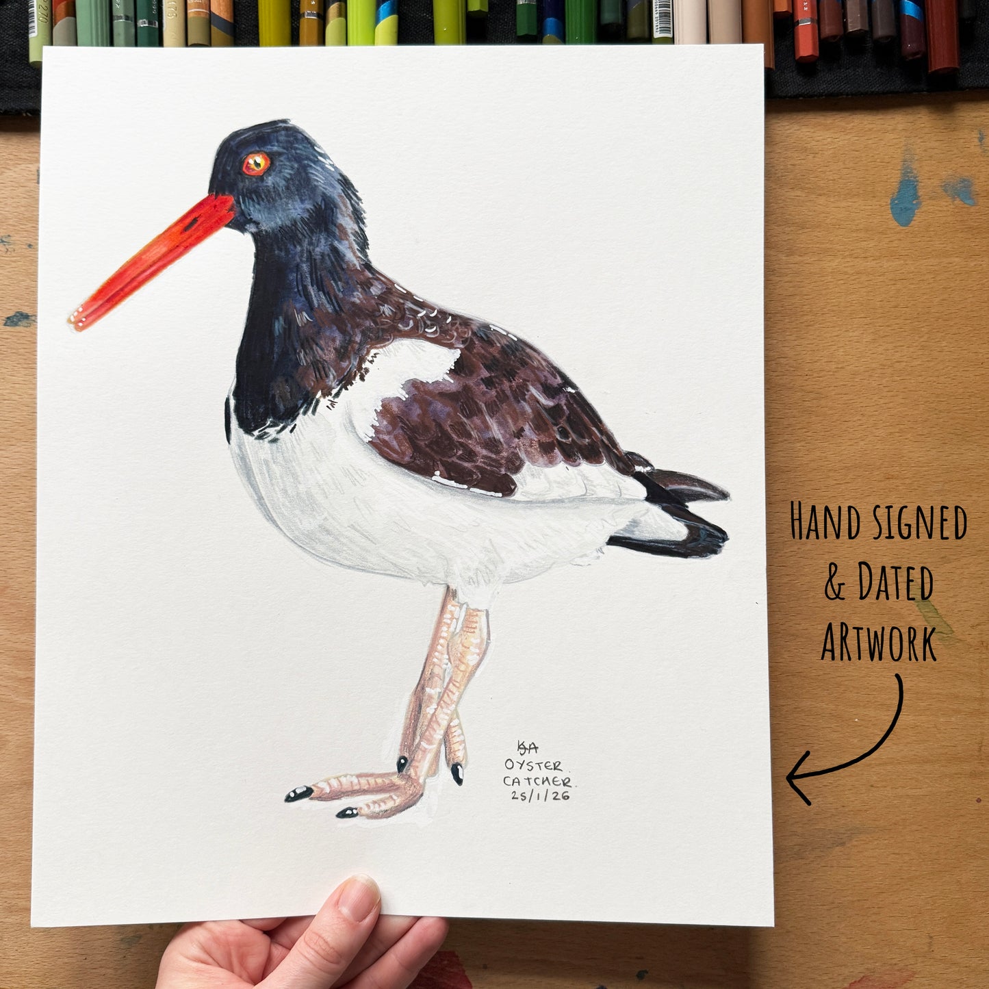 Hand-signed and dated artwork of a bird on a wooden surface with colored pencils in the background.
