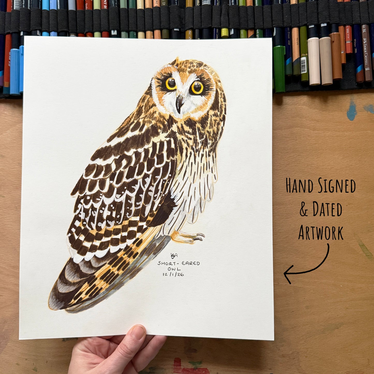 Hand-signed and dated artwork of an shorted eared owl on a white paper, held by a hand with art materials in the background.