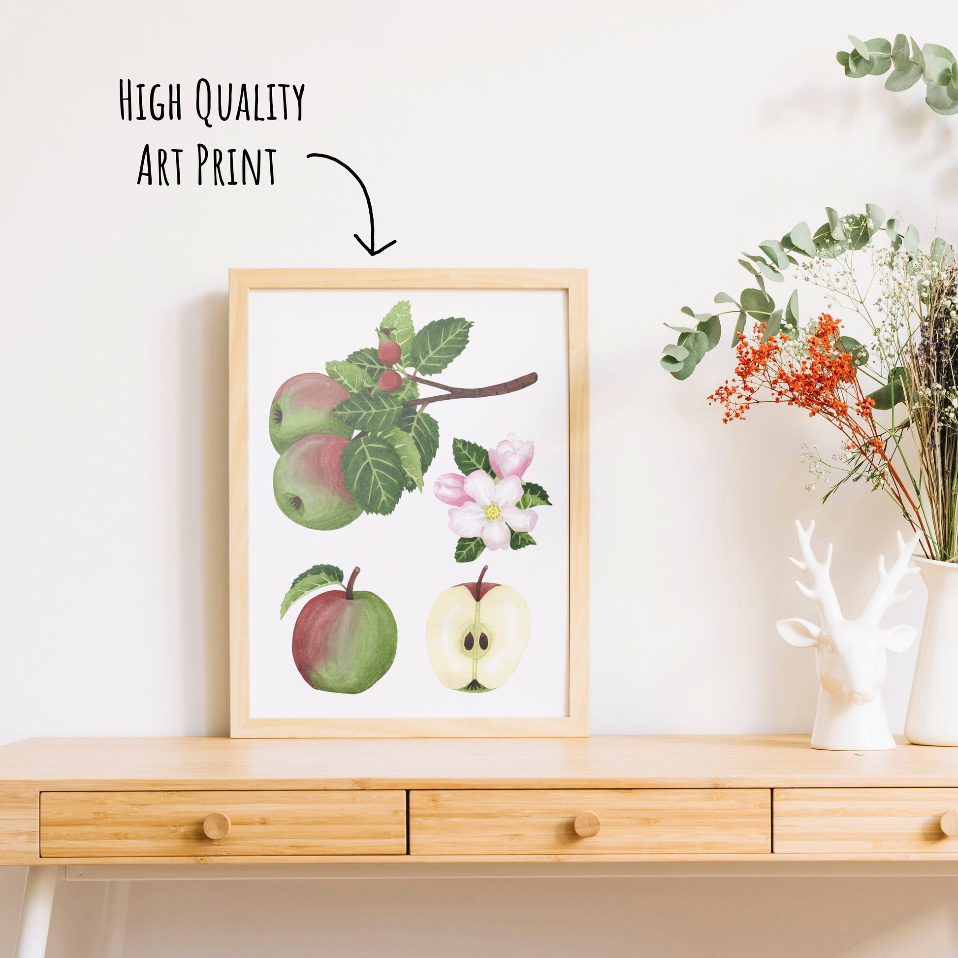 An apple study botanical art print shown in a frame on a table to with a vase with dried foliage in it. (framed not included with purchase)