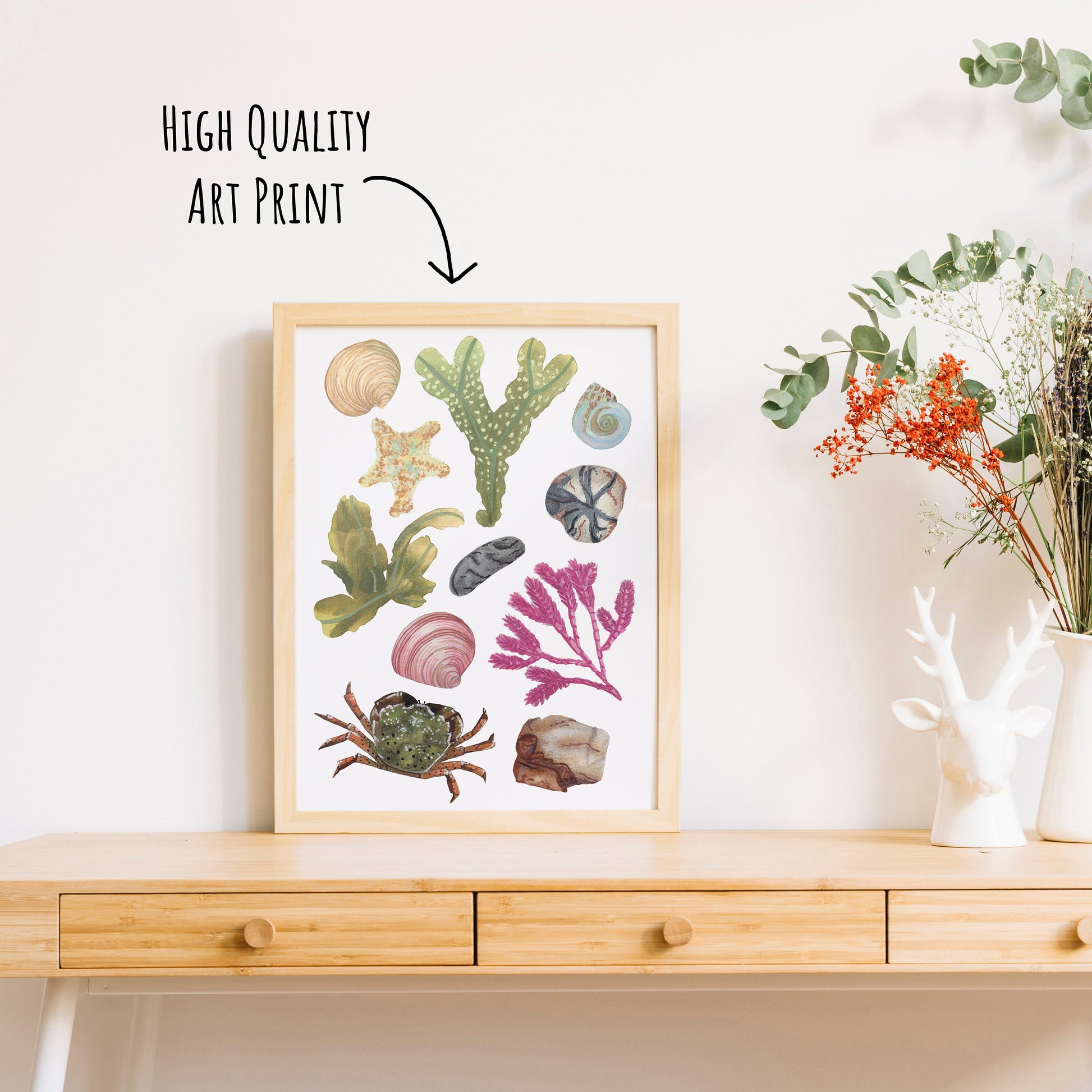 Showing an beach and rock pool wildlife art print in a frame on a table top with a vase of dried foliage. 