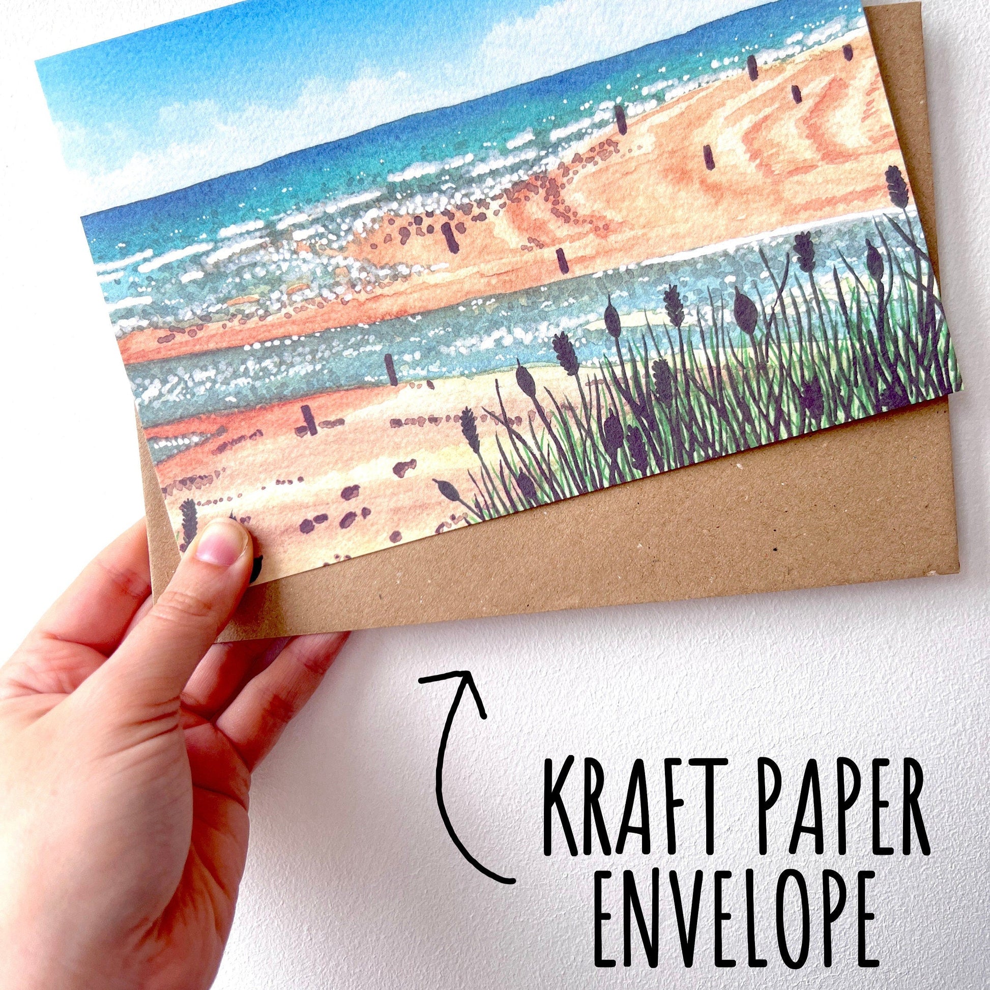 Hand holding a card with beach scene and kraft paper envelope on a white background with the font kraft paper envelope.