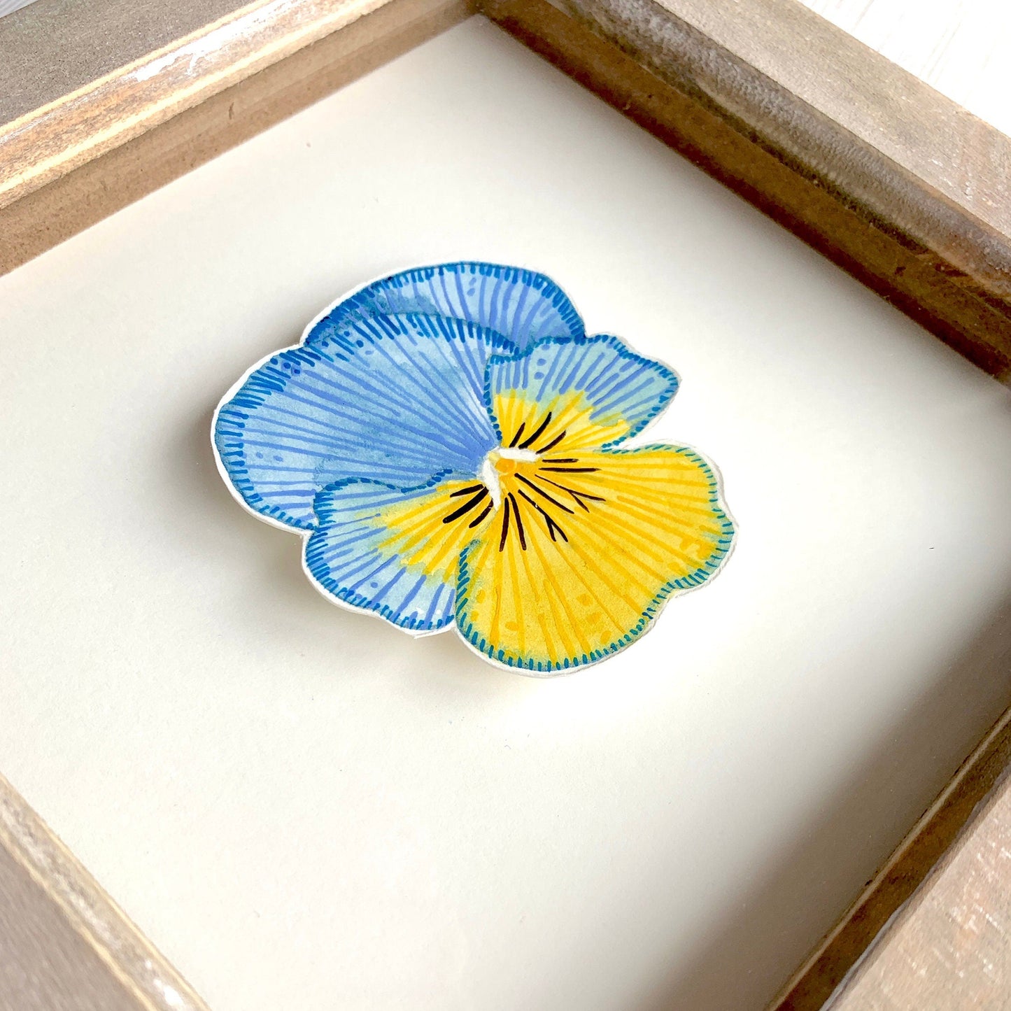 Framed artwork of a blue and yellow pansy flower on a smooth white paper background.
