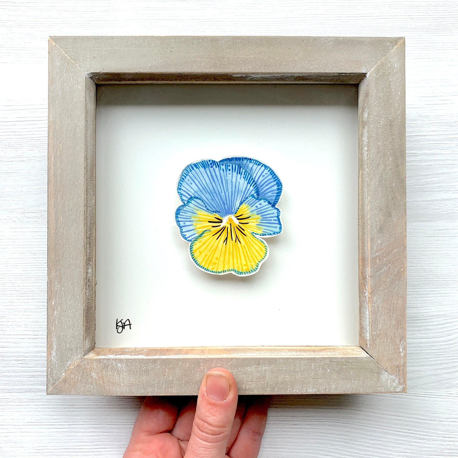 A painting of a blue and yellow pansy flower in a rustic wooden frame held up by a hand.