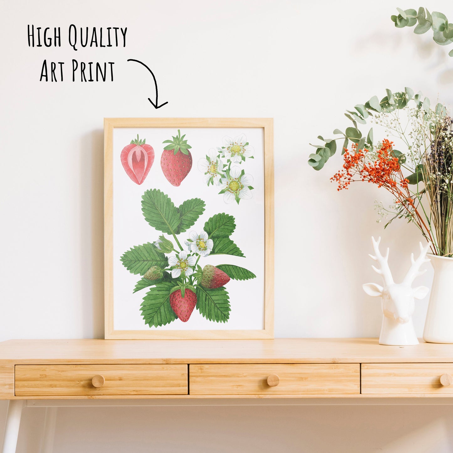 Botanical Strawberry Art Print in a light wooden frame on a table top with a vase of dried foliage.