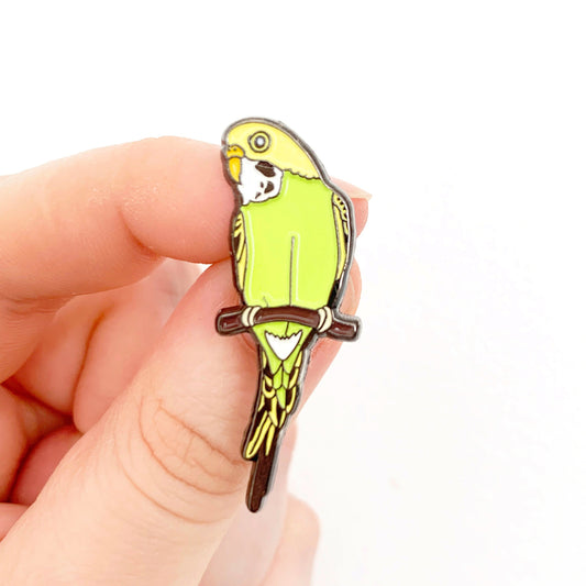 A hand holding a yellow and green budgie enamel pin badge. being held up against a white background.