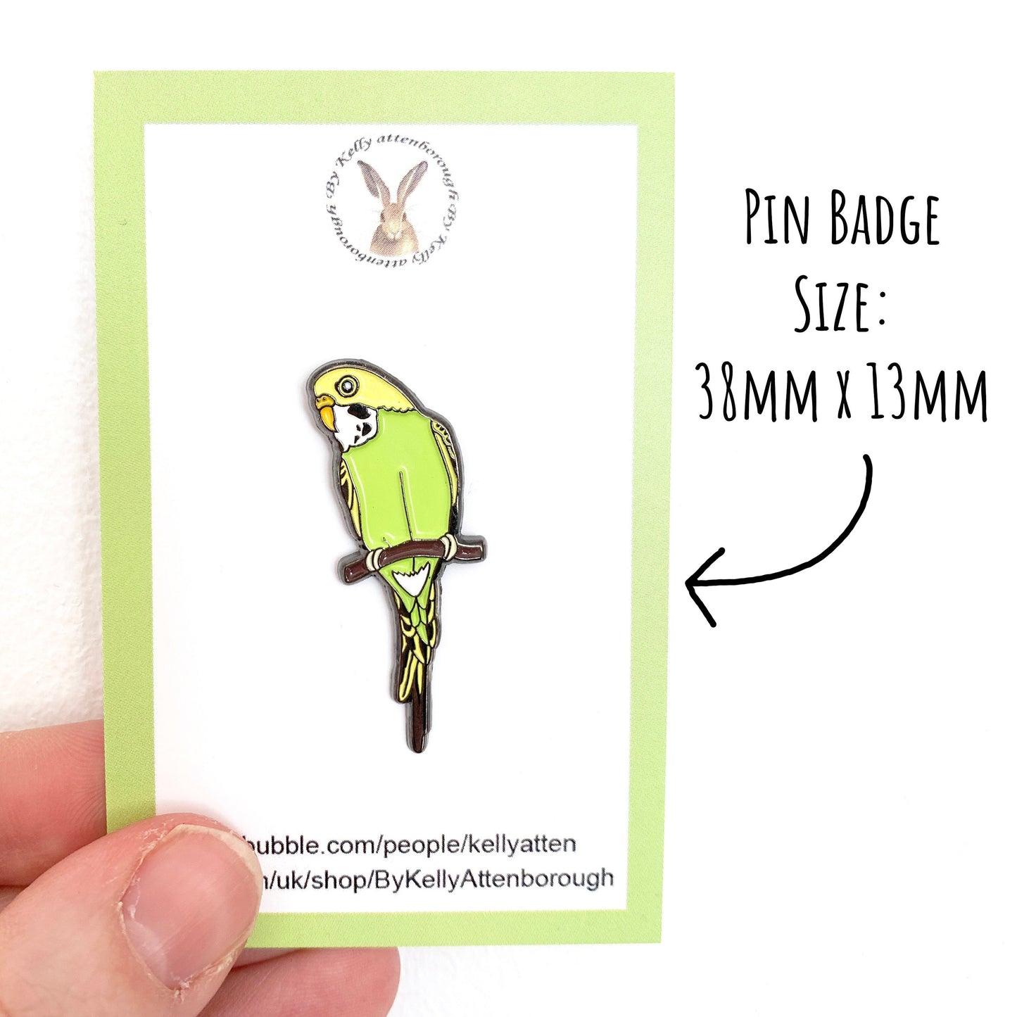 Budgie Enamel Pin Badge on a backing card. Being held up against a white background with the text pin badge size: 38mm x 13mm.