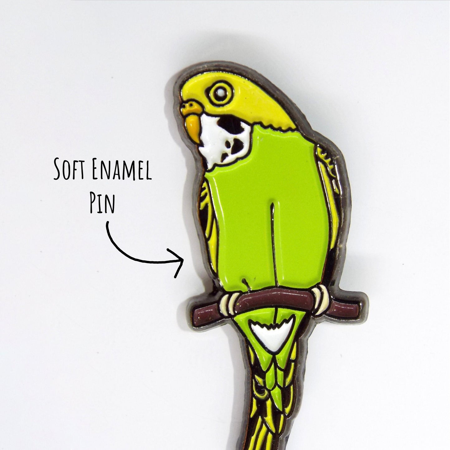 Budgie Enamel Pin Badge close up showing the details and textures of the pin badge. Pin laying on a white surface with the text soft enamel pin.
