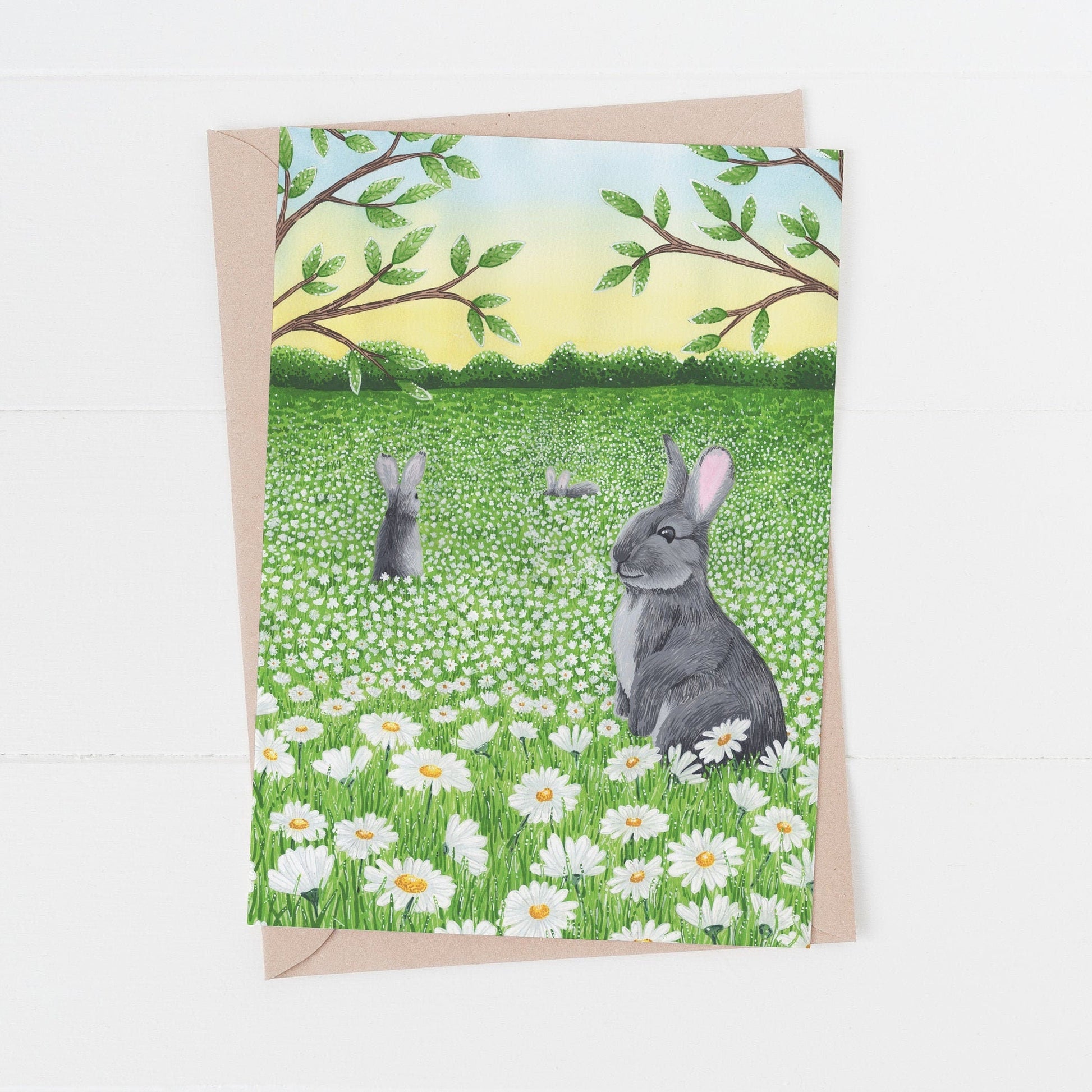 An illustrated Greeting card with two rabbits in a meadow of daisy flowers on a white background.