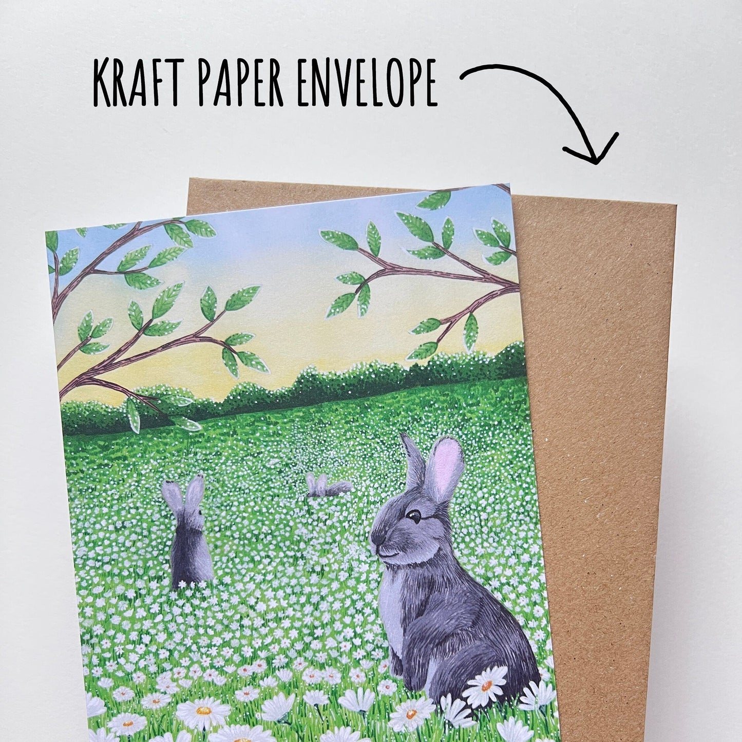 Daisy Meadow Rabbit Birthday Card bing held up with its kraft paper envelope against a white background. With Text kraft paper envelope.