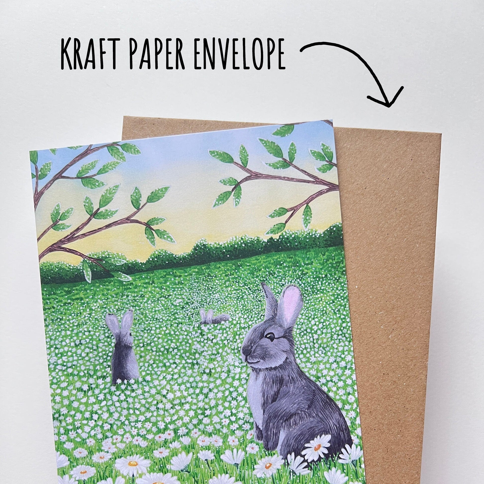 Daisy Meadow Rabbit Birthday Card bing held up with its kraft paper envelope against a white background. With Text kraft paper envelope.