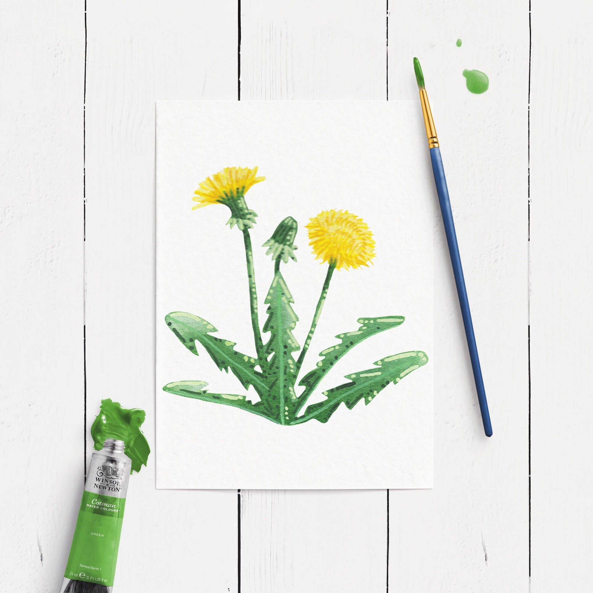 Watercolor painting of dandelions with a paintbrush and tube of green paint on a white wooden surface.