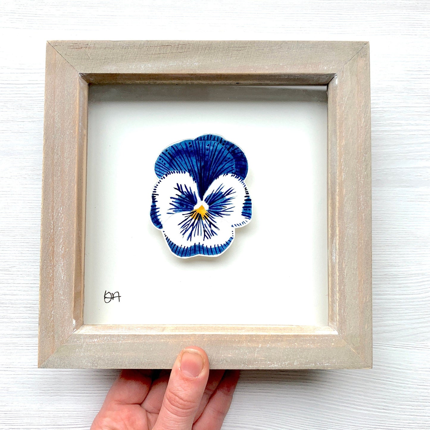 Framed artwork of a blue and white flower held by a hand on a light background