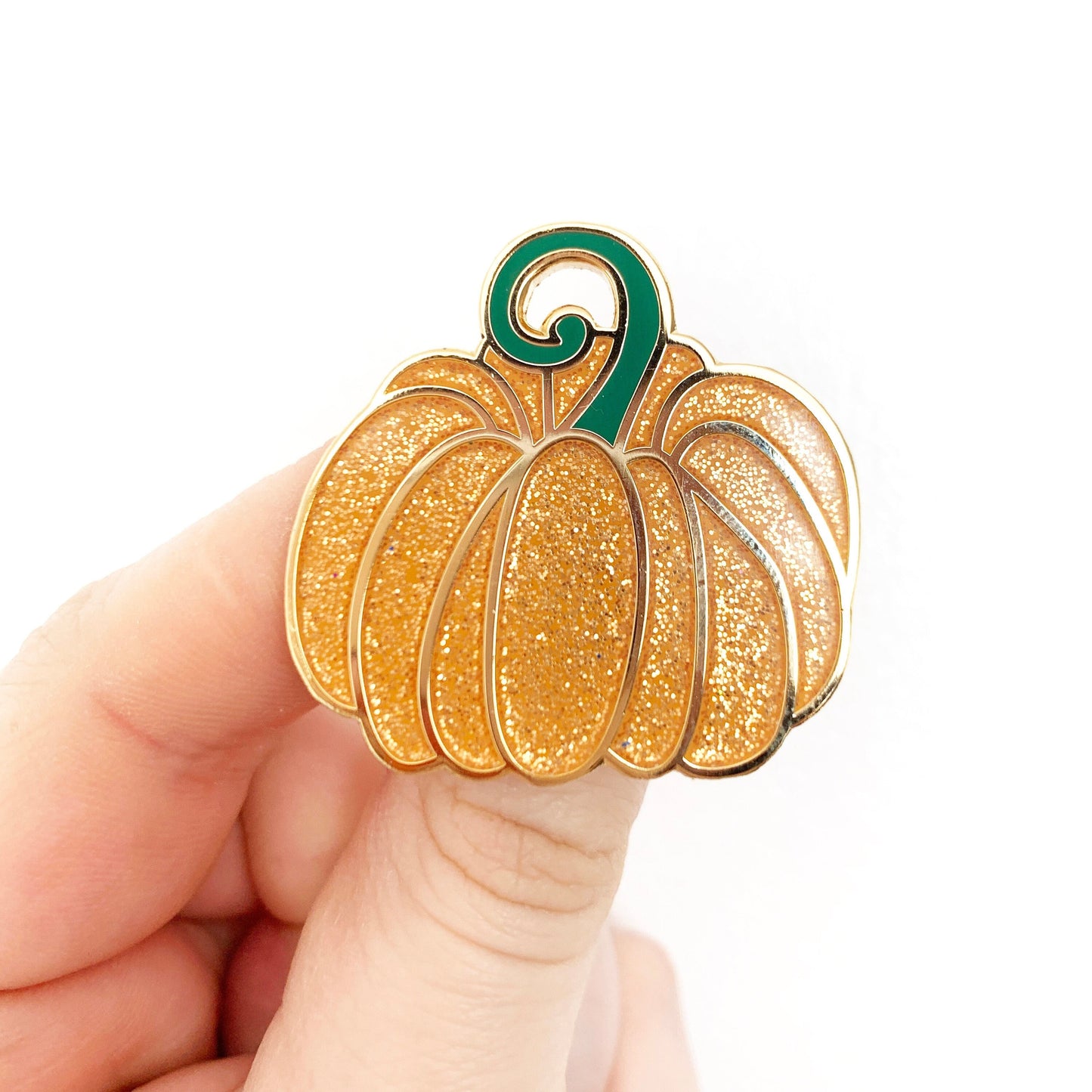 A hand holding a gold-coloured enamel pin shaped like a pumpkin with orange glitter and green enamel details.