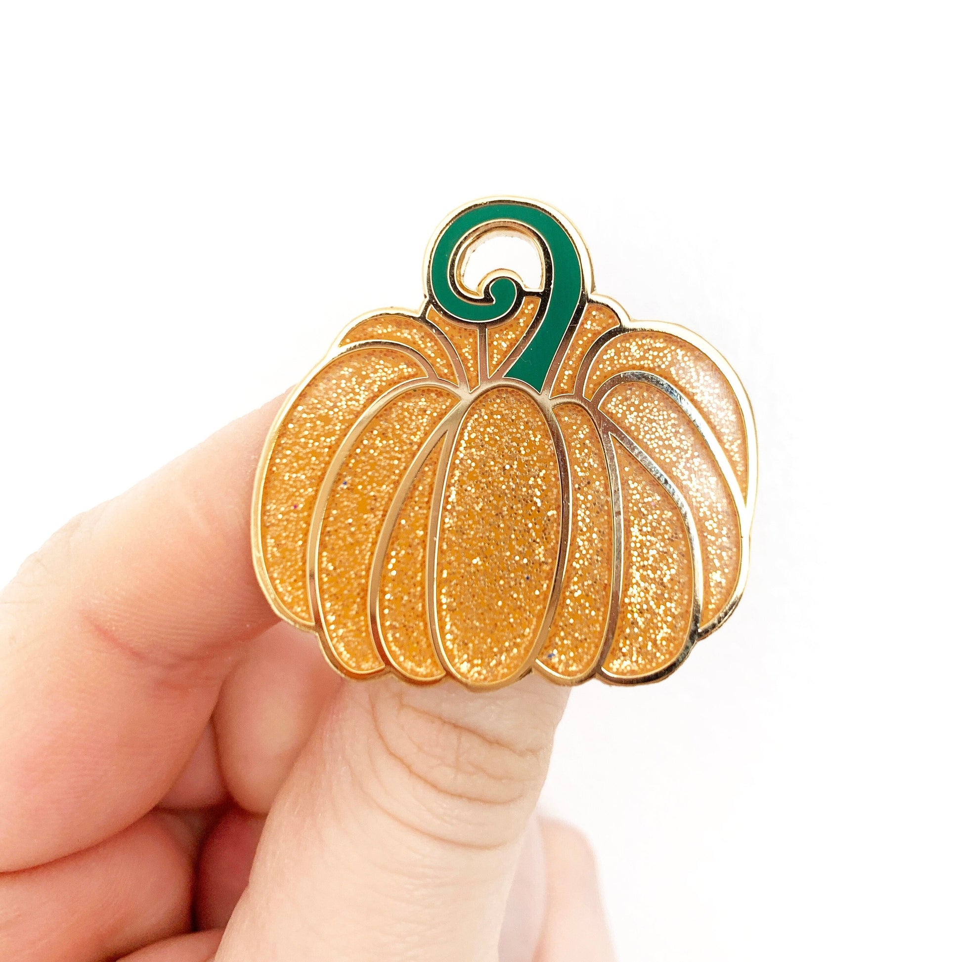 A hand holding a gold-coloured enamel pin shaped like a pumpkin with orange glitter and green enamel details.