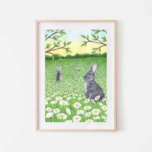 An art print featuring three rabbits in a field of daisy flowers with a bright blue sky and green grass, presented in a wooden frame.