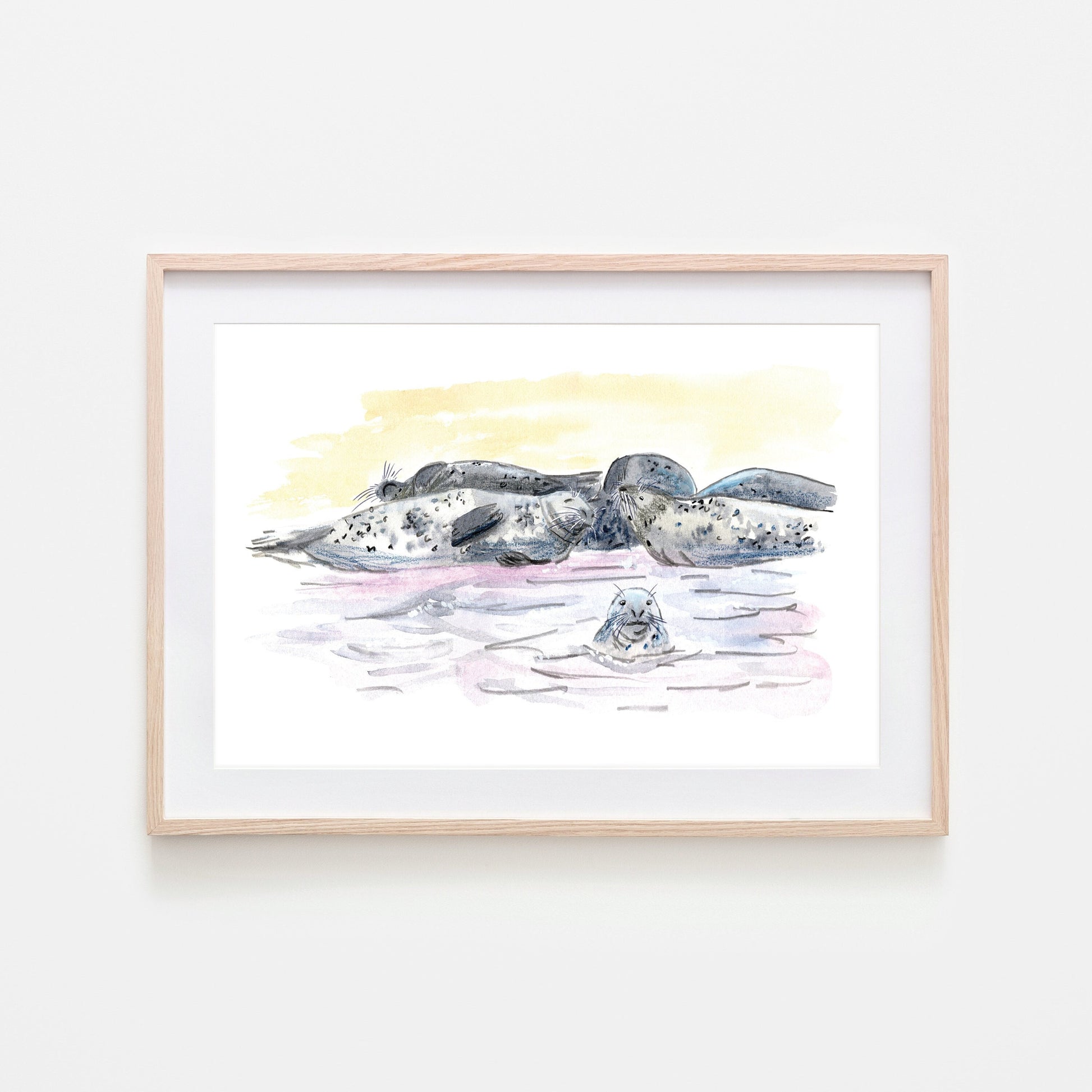 A framed art print depicting a group of grey seals resting on a beach, with watercolour and wax pastel techniques.