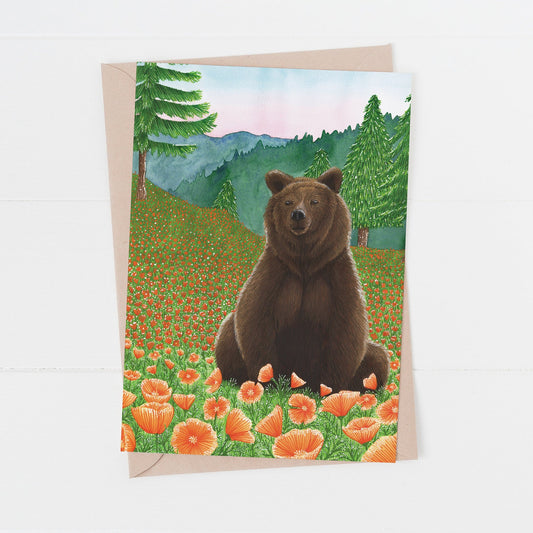 A greeting card featuring a watercolour illustration of a brown bear sitting in a field with orange poppies and a mountainous landscape in the background. Card laying on a white surface.