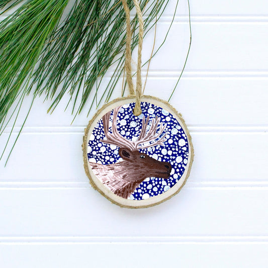 A hand-painted wooden Christmas ornament featuring a reindeer with a jute twine string for hanging, set against a dark blue background with snow.