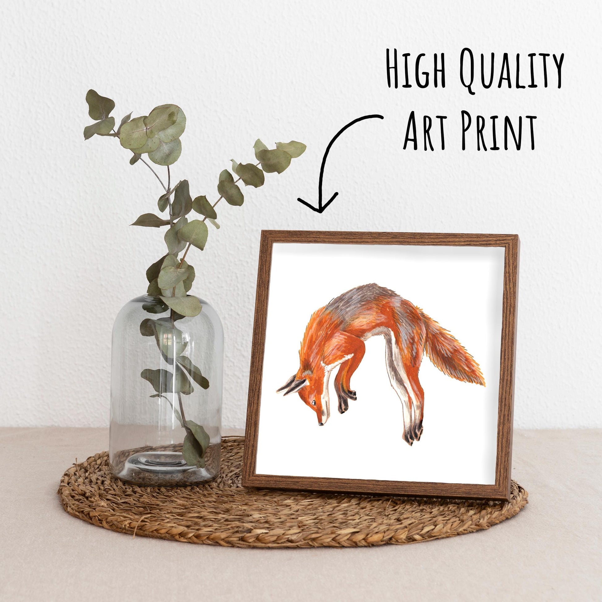 Jumping Red Fox Art Print framed in a dark wood frame on a straw placemat with a vase with a eucalyptus branch.