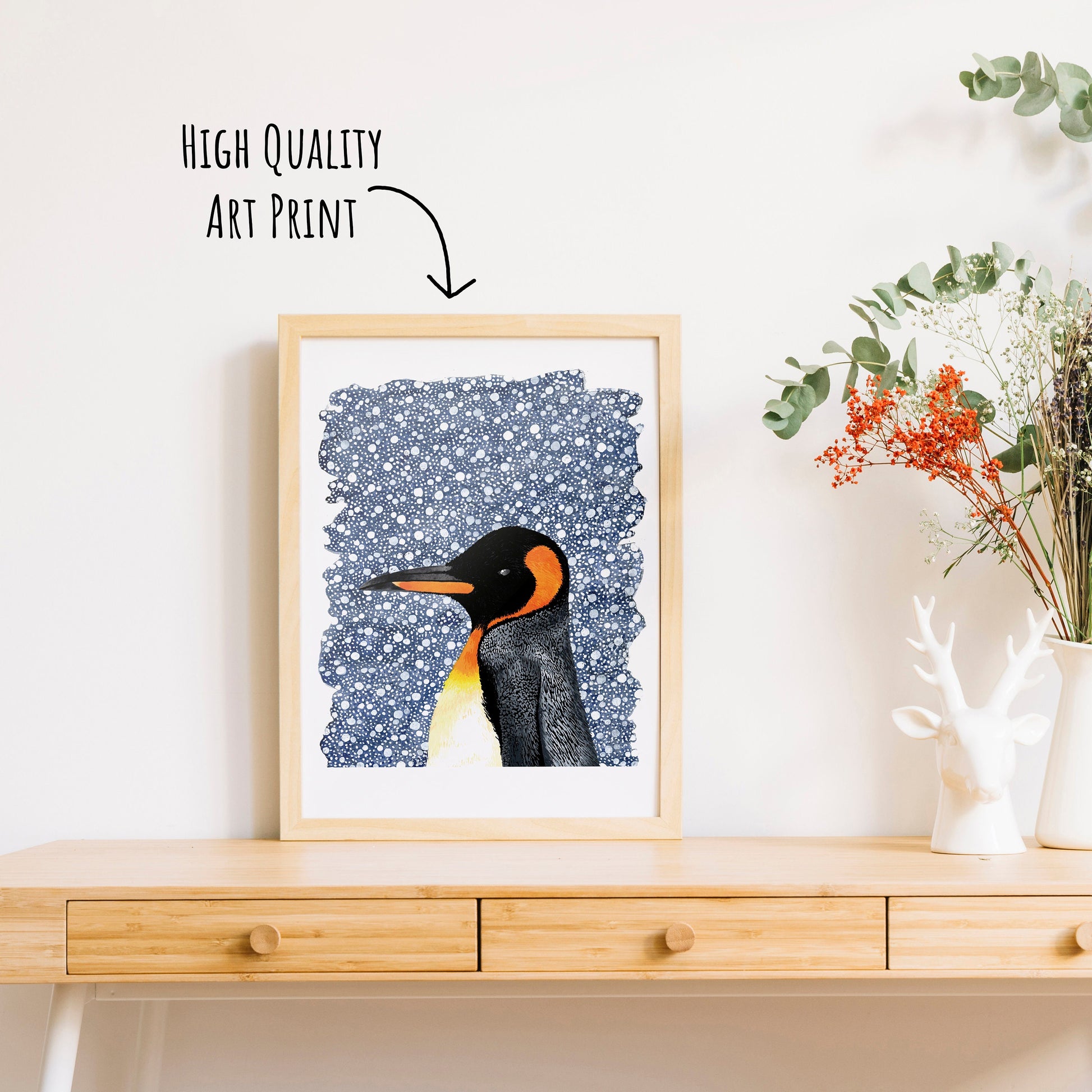 A king penguin with a deep blue snowy background art print in a frame on a table top with a vase full of dried foliage. (frame not included in purchase)