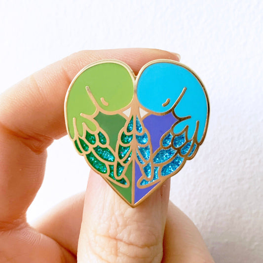 An enamel pin badge in the shape of a heart with a design of two love birds in green and blue colors with gold plating, held between fingers.