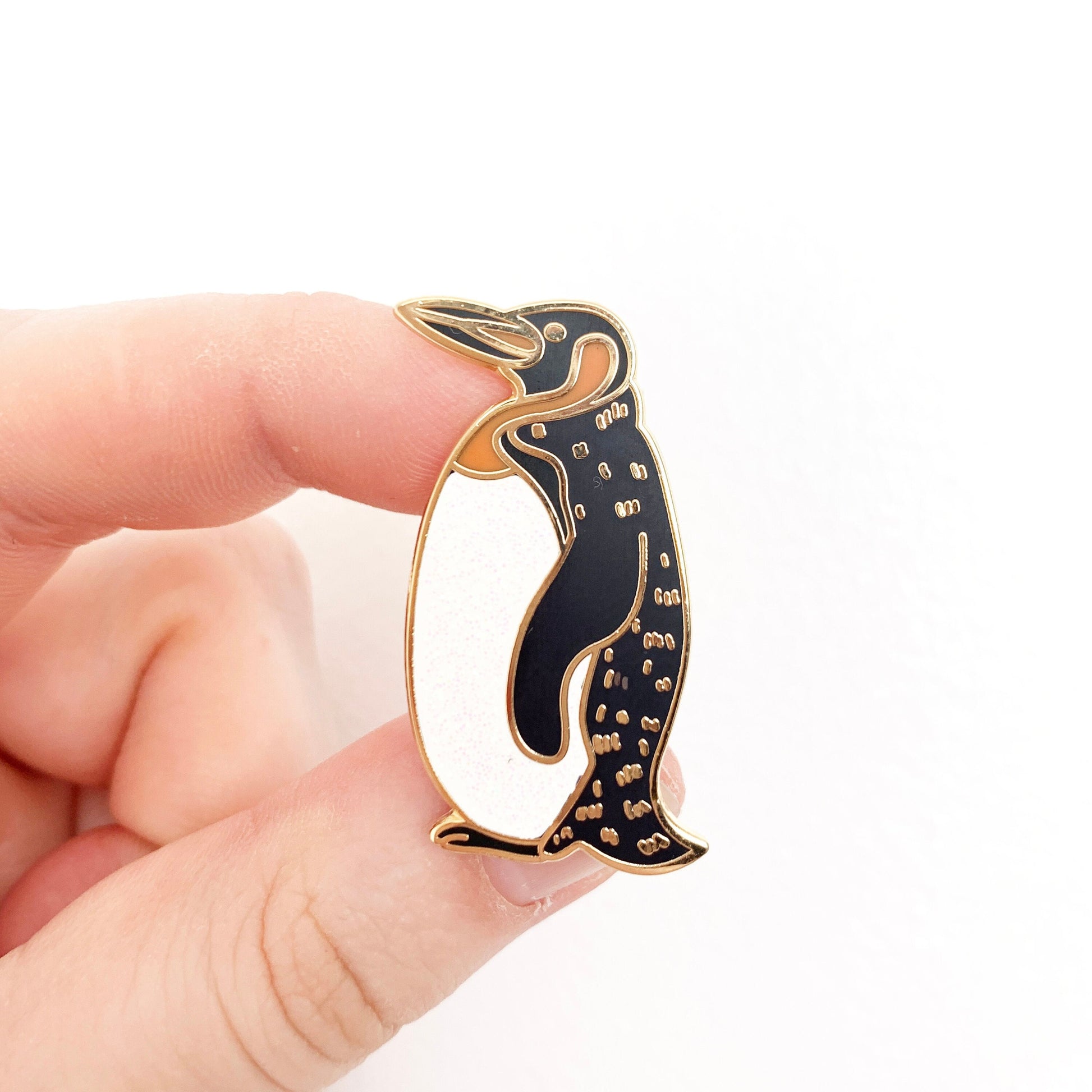A hand holding a black, white and gold penguin enamel pin badge, Against a white background.
