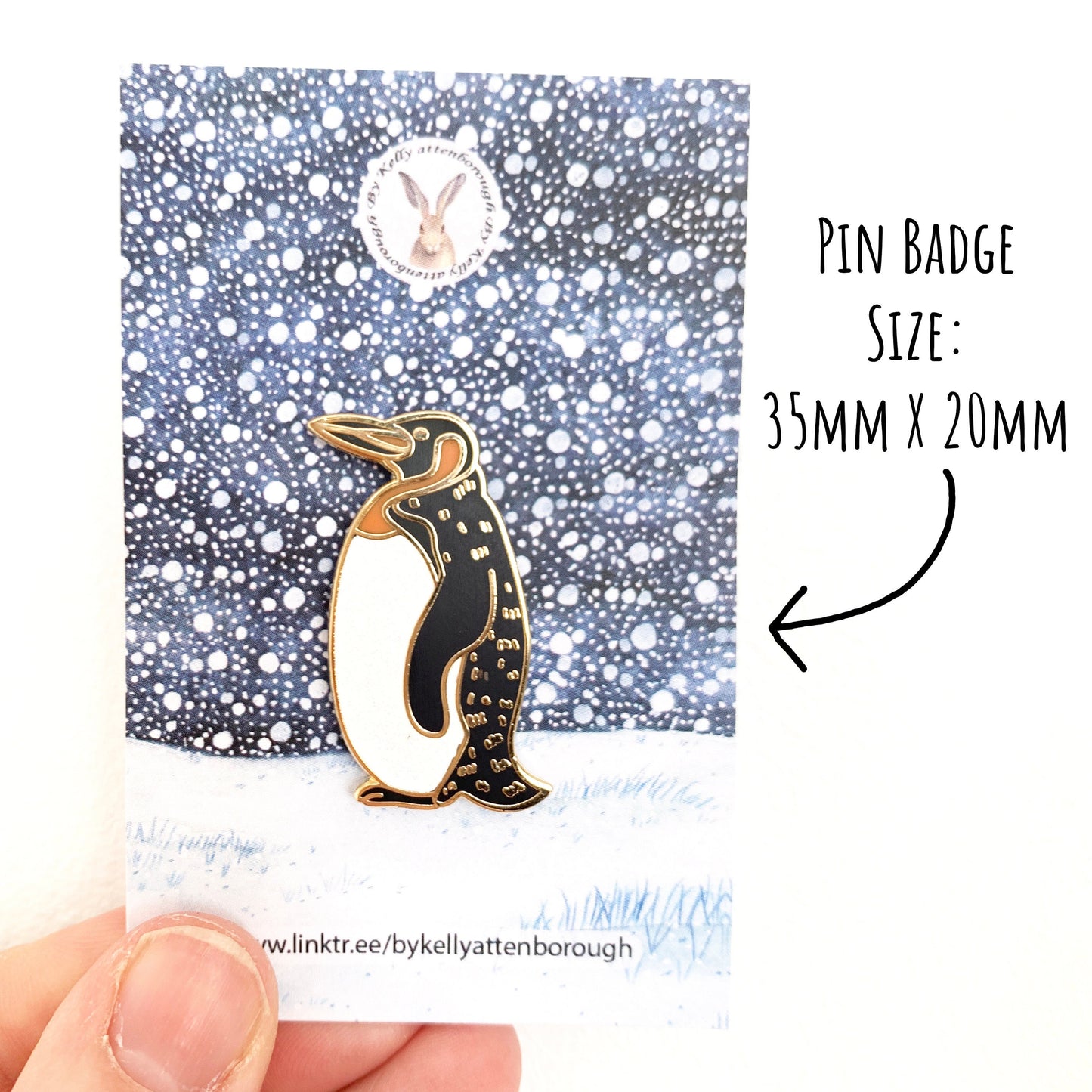 Penguin Enamel Pin Badge on its snowy scene backing card being held against a white background. With the text pin badge size: 35mm x 20mm.
