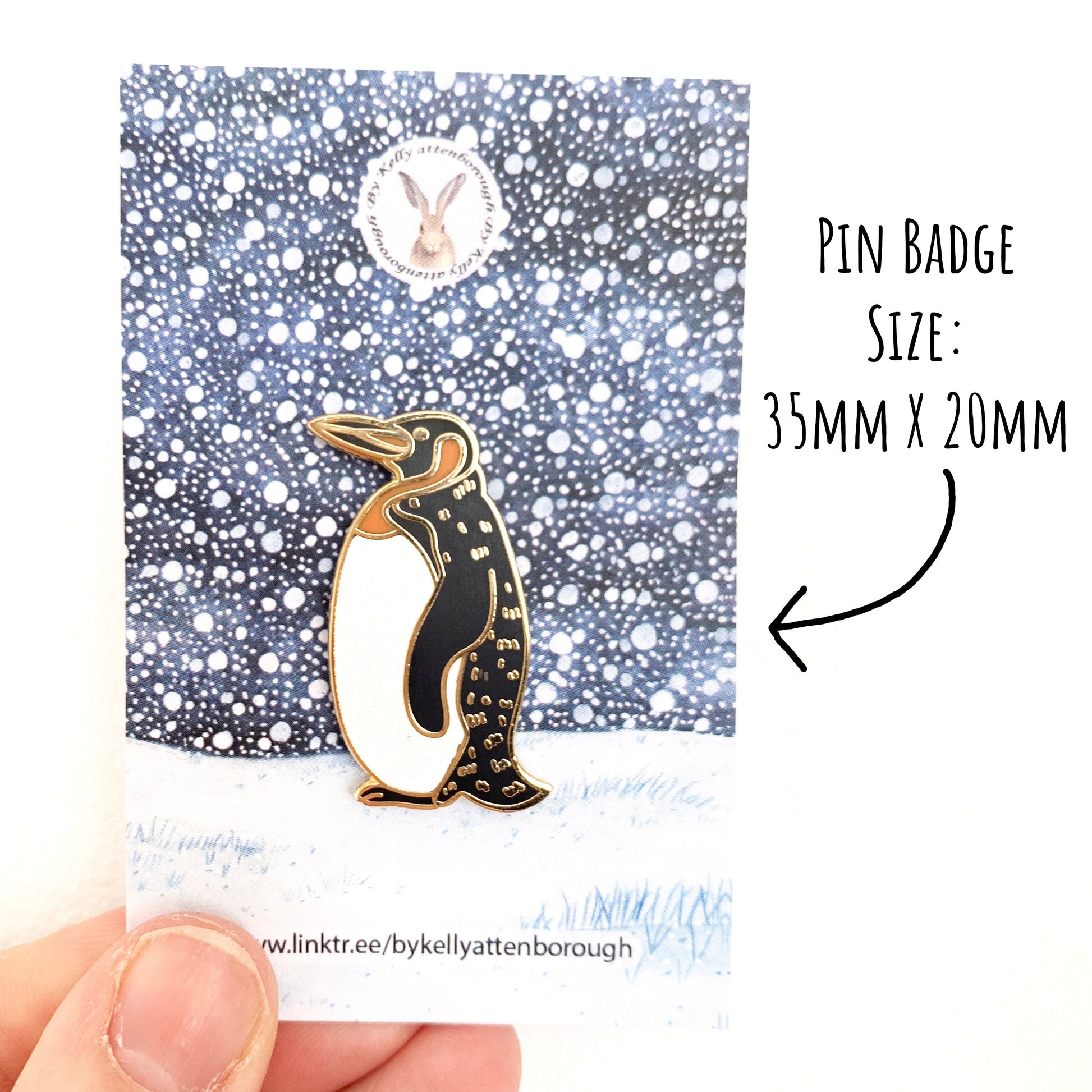 Penguin Enamel Pin Badge on its snowy scene backing card being held against a white background. With the text pin badge size: 35mm x 20mm.