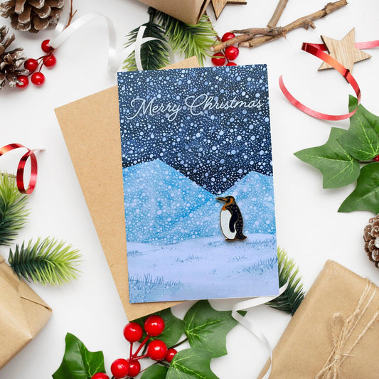 A Christmas card featuring a hand-painted snowy scene with a penguin on a dark blue background, accompanied by a 'Merry Christmas' message. The card is accompanied by a kraft paper envelope and festive decorations.