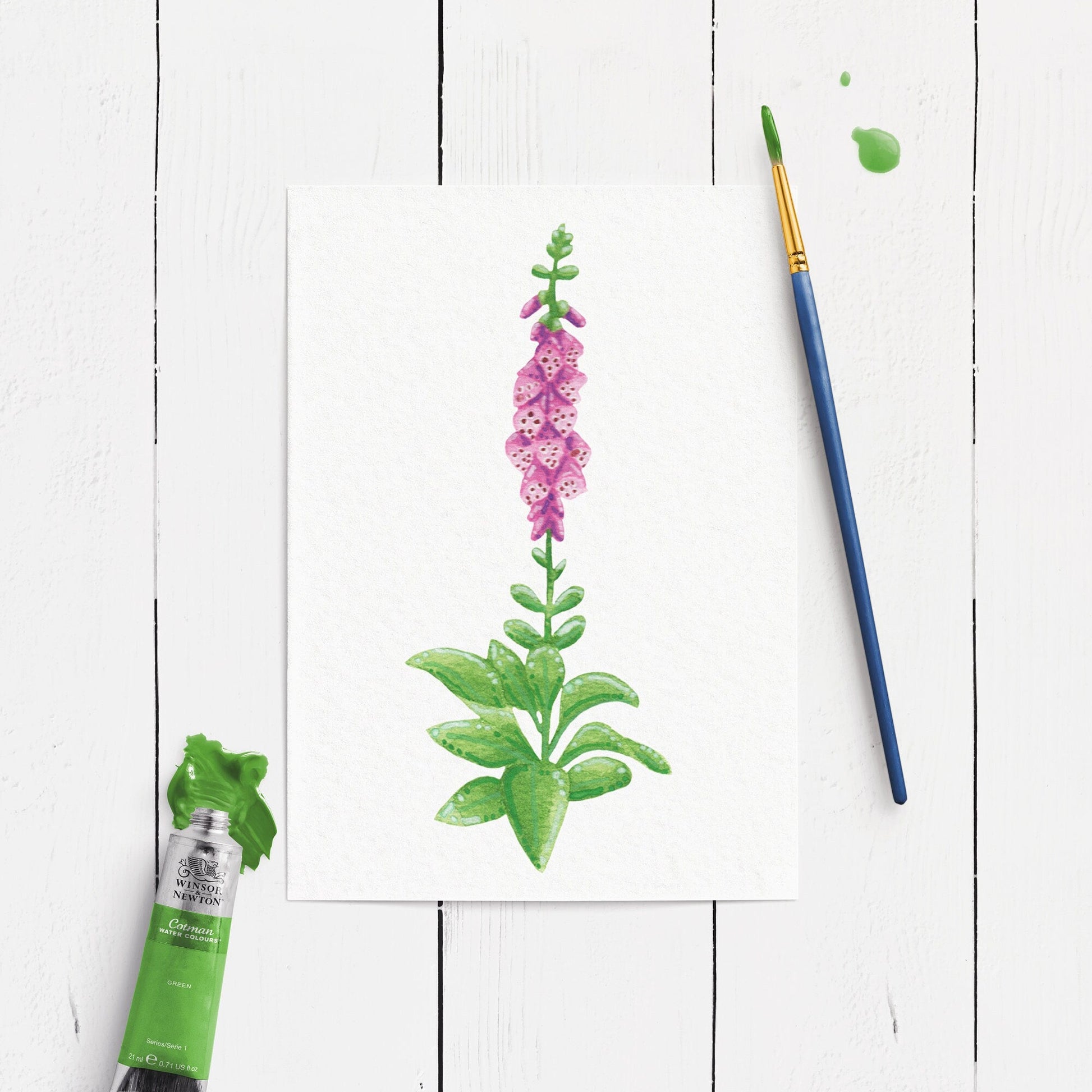 Painting of a pink flower foxglove with green leaves on a white wooden surface, accompanied by a paintbrush and paint.