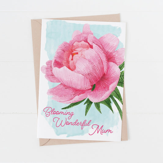A vibrant pink peony mothers day card with the text 'Blooming Wonderful Mum' on the front, displayed with its kraft paper envelope. Laying on a white surface.
