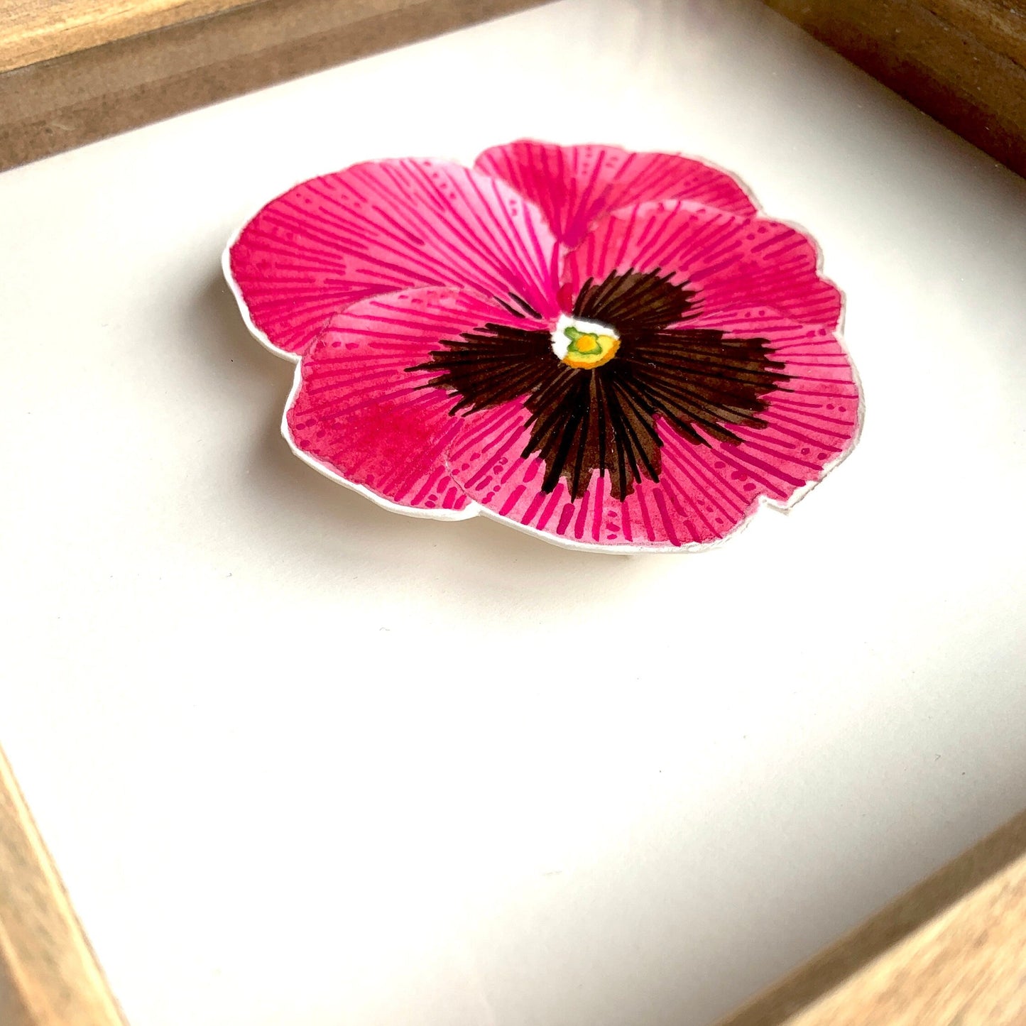 Pink pansy flower with a black center on a white card, framed in wood.